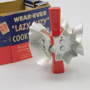 May include: A red plastic handle with multiple silver-colored metal cookie cutters attached in various shapes including a heart and a star. The tool is positioned in front of a blue and red cardboard box labeled Wear-Ever Lazy Suzy Cookie Cutter.