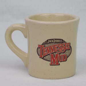 May include: Cream-colored ceramic mug with a curved handle. The mug features the text "Jack Daniel's Tennessee Mud" in a red and brown design. The mug has a speckled appearance.
