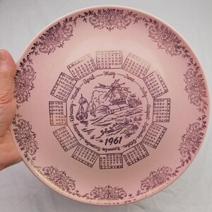 May include: A round pink ceramic plate featuring a dark purple 1961 calendar design. The center displays a windmill and sailboat illustration surrounded by twelve monthly grids. The border is decorated with intricate floral patterns in purple.