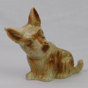 May include: A ceramic figurine of a dog in a sitting position, with its head tilted. The dog has a light green base color with brown markings on its ears, face, and body. The figurine has a small opening on its back.