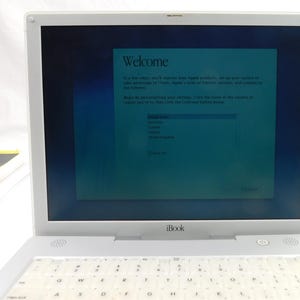 May include: A white iBook laptop with a keyboard and screen displaying a welcome message. The screen shows a list of countries. A book is visible in the background.