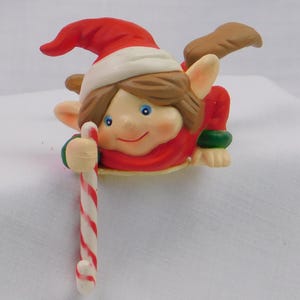 May include: A small elf figurine wearing a red hat with a white brim and a red and green outfit. The elf has brown hair, blue eyes, and pointed ears, and is holding a white and red striped candy cane in one hand while appearing to peek over an edge.
