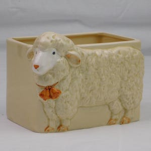 May include: Cream-colored ceramic planter with a sheep design. The sheep is sculpted in relief, with a fluffy, textured coat, a white face, and orange bells around its neck. The planter is rectangular with a smooth, matte finish.