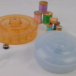 May include: An orange plastic thread organizer with a clear lid, a blue plastic container, and several spools of thread in various colors. The spools include pink, green, and purple thread. A single blue bobbin is also visible.