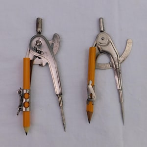 May include: Two vintage silver drafting compasses with attached yellow pencils. The compasses have a hinged design with a pointed tip and a pencil holder. One compass has a double-ring pencil holder.