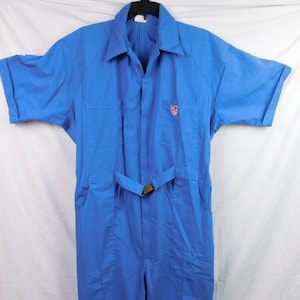 May include: A bright blue short-sleeved jumpsuit featuring a collared neckline, a front button-down closure, and two chest pockets. A small red and white shield-shaped patch is on the left chest pocket. An adjustable fabric belt with a metal buckle.