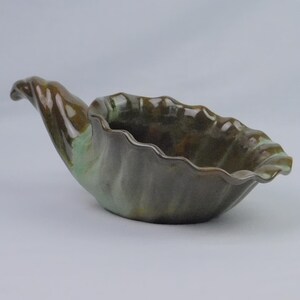 May include: A ceramic bowl shaped like a seashell with a glossy finish. The exterior and interior feature a blend of deep brown, earthy green, and muted teal tones. The rim has a wavy, scalloped edge, and the base tapers into a pointed handle.