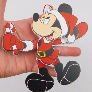May include: A wooden Christmas ornament featuring Mickey Mouse in a Santa Claus outfit. The ornament is red, black, and white, with a candy cane in one hand. The ornament is held by a hand.