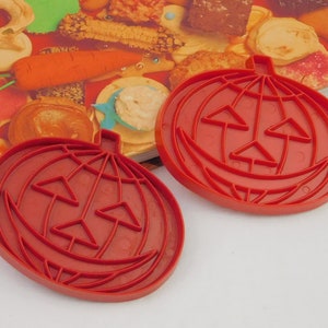 May include: Two red plastic pumpkin-shaped cookie cutters. Each cutter has a smiling jack-o'-lantern face design. The cutters are on a colorful book with images of cookies and other baked goods.