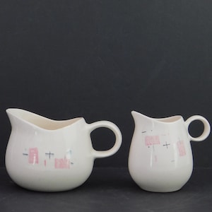 May include: Two off-white ceramic cream pitchers with a speckled finish. Each pitcher features a handle and is decorated with pink and blue geometric shapes. The pitchers have a mid-century modern design.