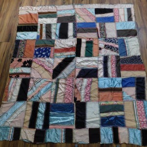 May include: A rectangular patchwork quilt constructed from various fabric scraps in diverse colors including black, blue, orange, pink, and white. The pieces are joined with visible zigzag stitching, creating a multi-textured surface.