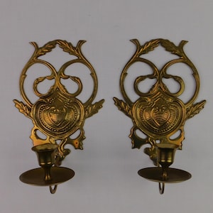 May include: A pair of matching brass wall sconces featuring an ornate, openwork backplate with leaf-like scrolls and a central shield-shaped motif. Each sconce includes a circular candle cup and a flat, round drip tray extending from the base.