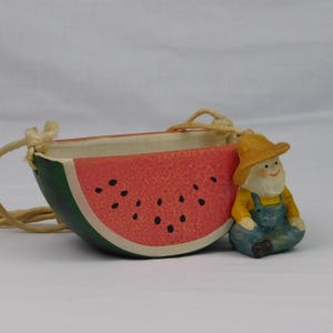 May include: A ceramic watermelon-shaped planter with a red interior, black seeds, and green rind. It has a beige rope for hanging. A small figurine of a farmer in overalls and a straw hat sits beside the planter. The planter is a decorative item.