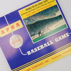 May include: A vintage APBA Professional Baseball Game box. The box is blue with yellow and white text. The box features a baseball graphic and a photo of a baseball game. Text includes "Manage the Big League Teams!" and "You are the Manager!"