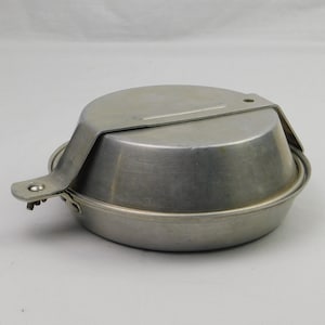 May include: A silver-colored camping mess kit with a lid and a handle. The kit appears to be made of metal and is designed for outdoor cooking and eating. The lid is attached with a metal bracket.
