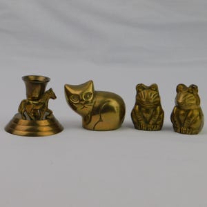 May include: A collection of brass figurines, including a horse-themed candle holder, a cat, and two frog figures. The objects are arranged on a white surface, showcasing their detailed craftsmanship and golden color.