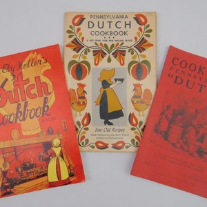 May include: Three vintage cookbooks with red and cream covers. The titles include "Pennsylvania Dutch Cookbook" and "Edna Ely Heller's Dutch Cookbook." The central book features a woman in traditional attire. The cookbooks are likely related to Pennsylvania Dutch cuisine.
