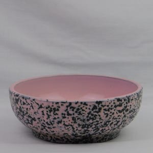 May include: A ceramic bowl with a speckled exterior and a solid pink interior. The bowl's exterior features a pattern of black speckles on a pink background. The bowl has a rounded shape and a slightly raised base.
