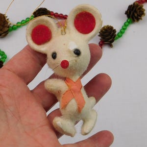 May include: A cream-colored felt mouse ornament with large ears featuring red circular centers, black bead eyes, a small red nose, and thin whiskers. The mouse wears a light orange ribbon tied around its neck and has a gold string for hanging.