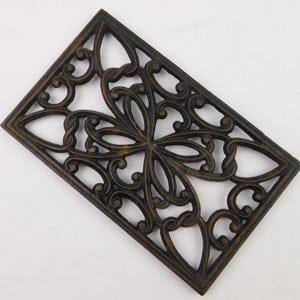 May include: A rectangular cast iron trivet featuring an intricate openwork design of symmetrical scrolling patterns and curved lines. The metal has a dark brown, weathered finish with subtle highlights, showcasing a decorative lattice structure.