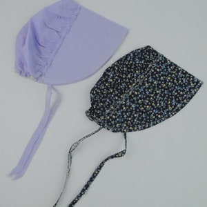 May include: Two sun hats with ties. One is a light purple sun hat with a ruffled edge. The other is a black sun hat with a white floral pattern.
