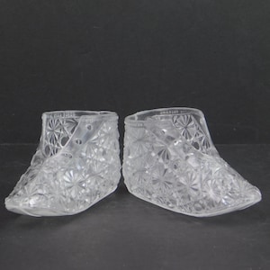 May include: A pair of clear glass shoe-shaped objects with a starburst pattern. The shoes have a lace-up design and the text "FOR CHILDREN" is visible on the top edge. The shoes are likely decorative or collectible items.