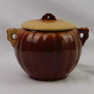 May include: A round, glazed ceramic bean pot with a dark brown, ribbed body and two side handles. The pot features a flat, light tan lid with a small central knob that has a hole in the middle. The overall design is bulbous and symmetrical.
