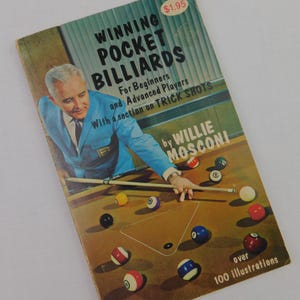 May include: Vintage book cover titled "Winning Pocket Billiards" featuring a man in a blue suit aiming a pool cue. The cover includes text about trick shots and illustrations, with a price tag of $1.95.