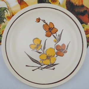 May include: A round off-white ceramic plate featuring a hand-painted floral design in shades of yellow and orange with brown stems and leaves. The plate has a thin brown circular rim accent along the edge, providing a simple and clean aesthetic.