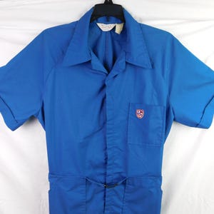 May include: A bright blue short-sleeved jumpsuit featuring a collared neckline, a button-down front, and a single chest pocket adorned with a small, colorful shield-shaped emblem. The garment includes cuffed sleeves and a waist adjustment buckle.
