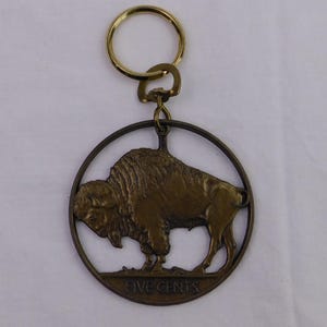 May include: A bronze-colored key ring featuring a circular pendant with a detailed depiction of a bison. The pendant has the words "FIVE CENTS" at the bottom. The key ring includes a gold-colored split ring.