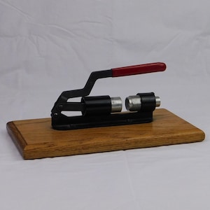 May include: A black metal crimping tool with a red handle, mounted on a wooden base. The tool has a lever design and is used for joining or securing items. The wooden base is rectangular and has a natural wood grain.