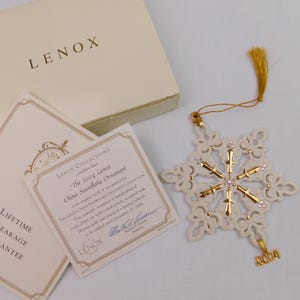 May include: A Lenox 2004 china snowflake ornament with gold accents and crystal jewels, featuring a gold tassel and a 2004 charm. The set includes a branded box and a certificate of authenticity stating it is crafted with 24 karat gold.