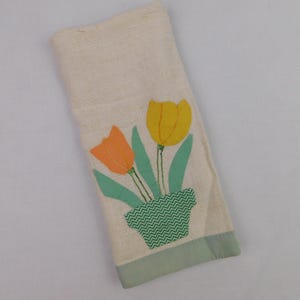 May include: A cream-colored hand towel with an appliqued design of two tulips in a green zig-zag patterned pot. One tulip is orange, the other is yellow, with green leaves. The bottom edge has a light green border.