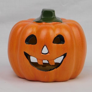 May include: A ceramic jack-o'-lantern pumpkin figurine with a bright orange exterior, a dark green stem, and cutouts for the eyes, nose, and a wide, toothy grin. The surface features subtle vertical ridges and a smooth, glossy finish.