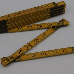 May include: A vintage wooden folding ruler, with black joints and yellow painted wood. The ruler is marked with inch measurements and numbers. The ruler is partially folded and unfolded, showing the measurement markings.