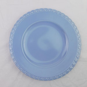 May include: A round, light blue ceramic plate featuring a smooth, flat center and a raised, textured border with a continuous rope-like pattern around the entire outer edge. The plate has a uniform, solid color finish throughout its surface.