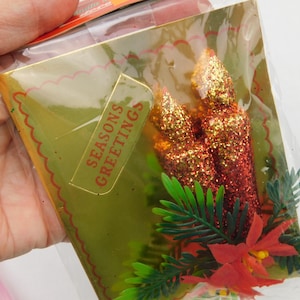 May include: A rectangular gold-toned card featuring the text SEASONS GREETINGS, adorned with two glittery red candle-shaped ornaments, green plastic pine needles, and a red poinsettia flower, all enclosed in a clear plastic protective sleeve.