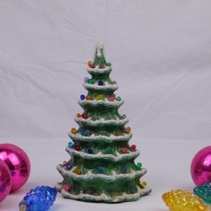 May include: A green ceramic Christmas tree with white accents and colorful lights. The tree is surrounded by pink ornaments and blue and yellow decorative pinecone-shaped lights, creating a festive holiday scene.