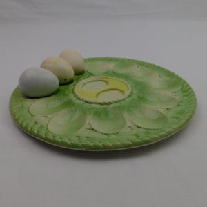 May include: A green ceramic deviled egg serving platter with a decorative leaf design and a braided edge. Three eggs are placed on the platter. The center of the platter has a yellow and white design.