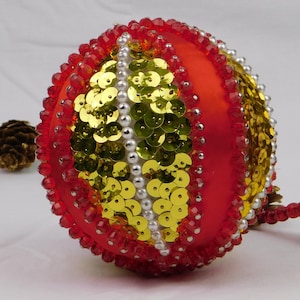 May include: A spherical ornament featuring a red satin base decorated with vertical rows of gold sequins, white pearl-like beads, and faceted red beads. The design is segmented into panels, creating a textured, multi-layered appearance.