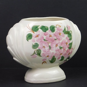 May include: A white ceramic vase with a wide, open top and a pedestal base. The vase is decorated with a cluster of pink flowers and green leaves. The vase has a shell-like design on the sides.
