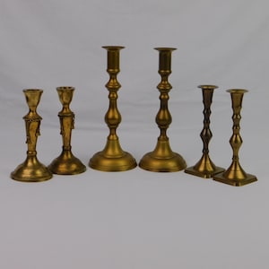 May include: A collection of six brass candlesticks in varying heights and designs. The candlesticks have a gold finish and feature ornate details, including fluted stems and decorative bases. The arrangement includes both tall and short candlesticks.