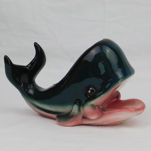 May include: A ceramic whale figurine with a glossy finish. The whale is primarily dark teal with a pink and white mouth. The tail fins are black. The whale is open-mouthed, and the interior of the mouth is pink.