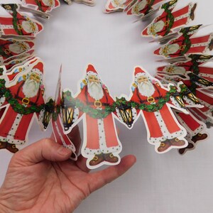 May include: A paper garland featuring a repeating die-cut design of Santa Claus figures. Each Santa wears a red suit with white fur trim, holds a black lantern, and is connected to the next figure by a green holly garland across the waist.