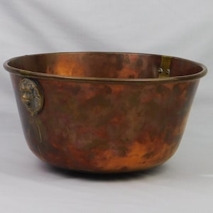 May include: A copper bowl with a rounded shape and a bronze rim. A bronze lion's head is affixed to the side. The bowl has a mottled, aged appearance with shades of brown and orange.