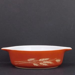 May include: An oval-shaped casserole dish with a vibrant orange exterior and a white interior. The exterior features a tan-colored wheat stalk pattern printed horizontally across the center. The dish includes two integrated handles on each side.