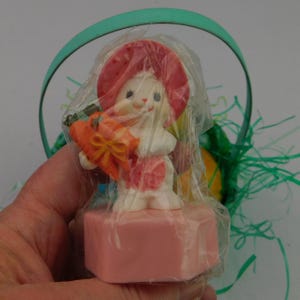 May include: A white bunny figurine wearing a pink hat and dress, holding a carrot with an orange bow. The bunny is on a pink base, wrapped in plastic, with a green basket handle in the background. Easter-themed.