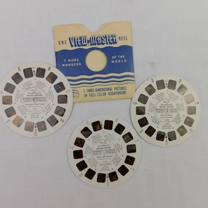 May include: Three vintage View-Master reels and a blue and white sleeve. The reels are white with small rectangular windows containing images. The sleeve reads "ONE VIEW-MASTER REEL" and "7 MORE WONDERS OF THE WORLD".