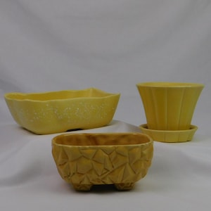 May include: Three yellow ceramic planters of varying shapes. One is a long, boat-shaped planter with a speckled finish. Another is a geometric, rectangular planter with a textured surface. The third is a flared planter with a matching saucer.